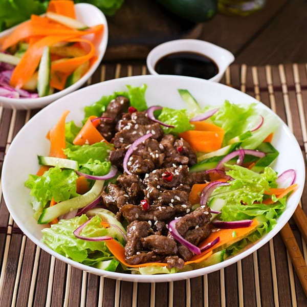 Asian beef salad with fresh vegetables, sesame seeds, and chili in a white bowl.