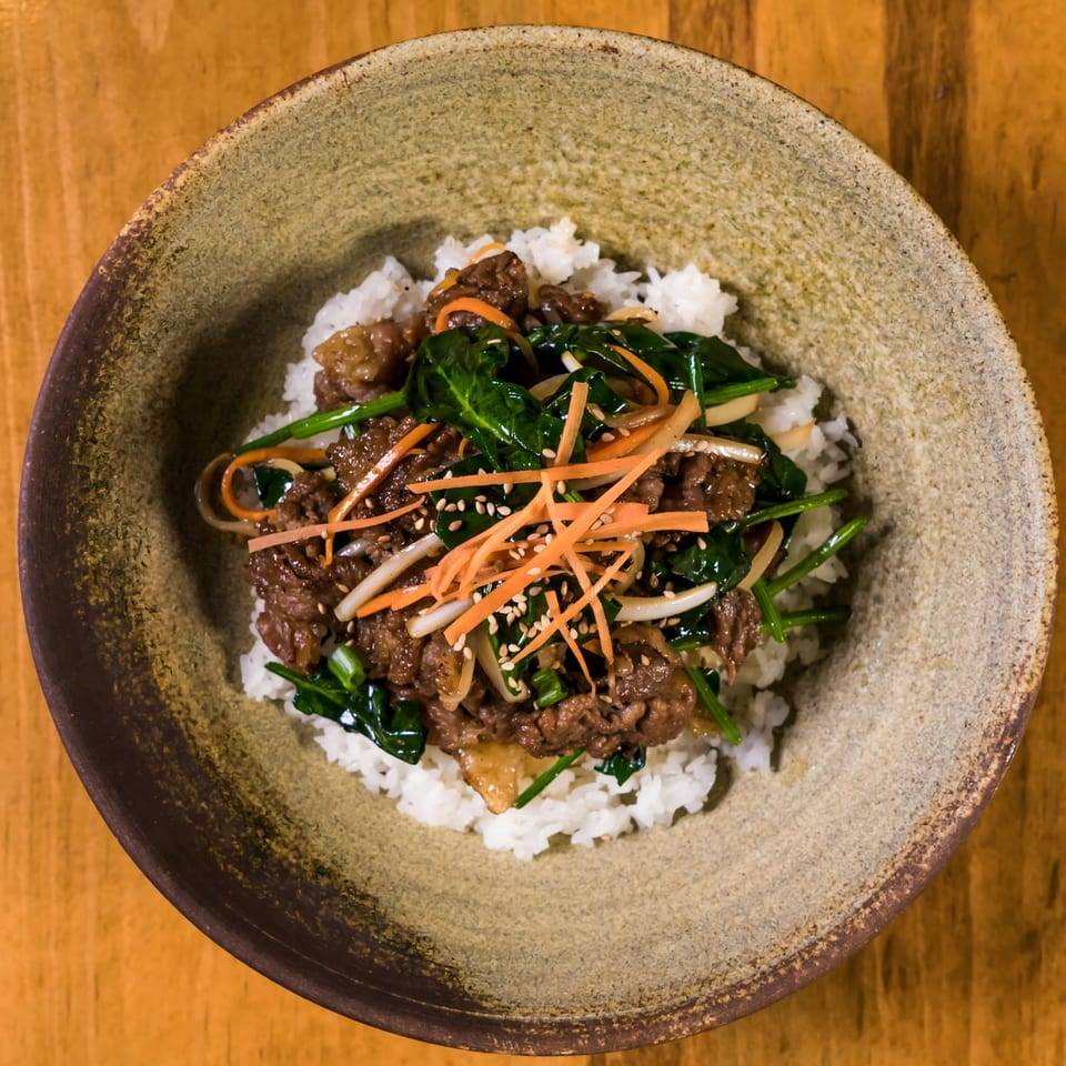 Asian beef and spinach stir-fry with shredded carrots and sesame seeds over white rice in a bowl.