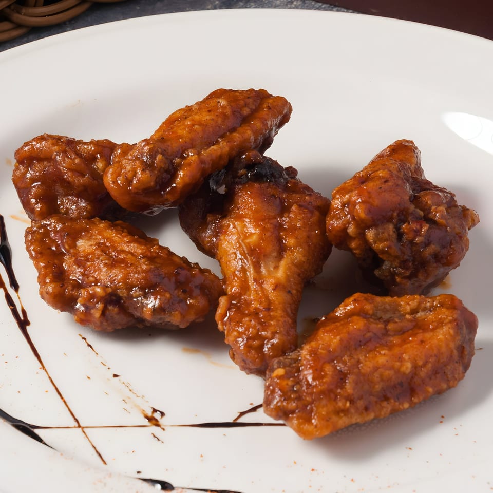 Six Asian-style chicken wings coated in a glistening brown sauce, artfully arranged on a white plate.